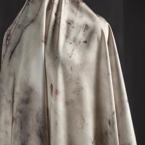 May include: A draped fabric with a marbled appearance. The off-white material is marked with dark gray and reddish-brown blotches, creating a textured, organic pattern. The fabric is draped over an unseen form, with soft folds.