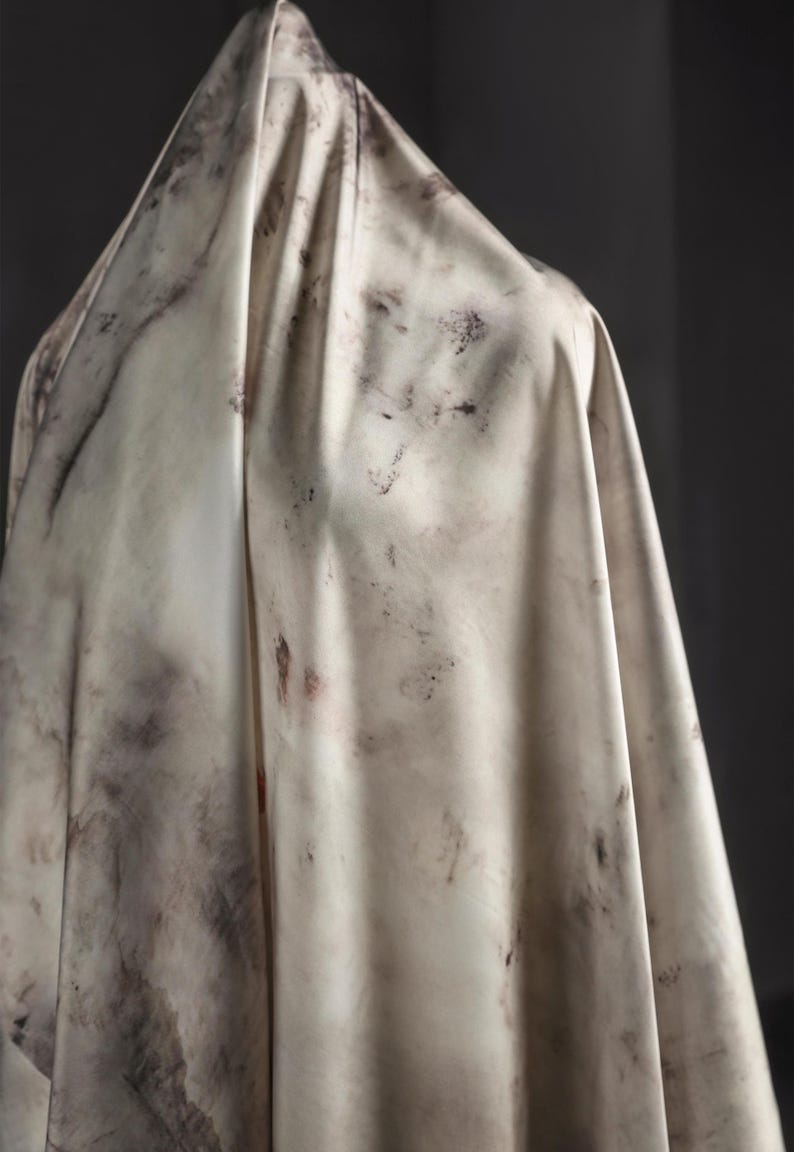 May include: A draped fabric with a marbled appearance. The off-white material is marked with dark gray and reddish-brown blotches, creating a textured, organic pattern. The fabric is draped over an unseen form, with soft folds.
