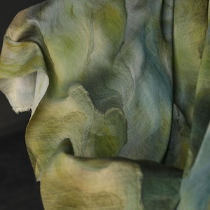 Puede incluir: Primer plano de una tela fluida con un patr&oacute;n similar a una acuarela. La tela presenta tonos de verde, azul y beige, creando un efecto marmolado. El material parece ligero y semitransparente, con textura y pliegues visibles.