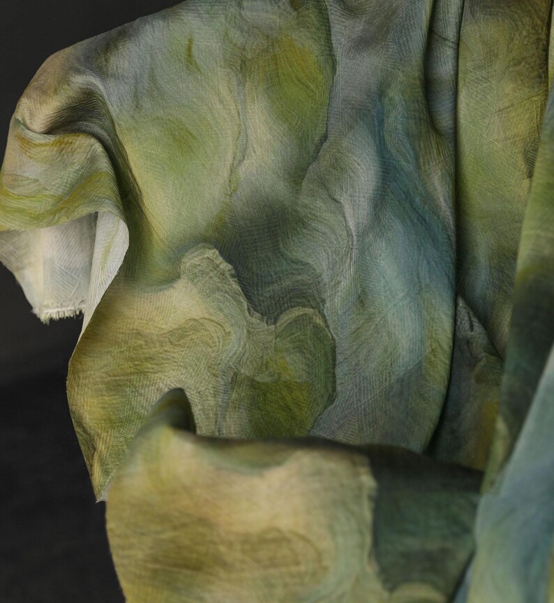 Puede incluir: Primer plano de una tela fluida con un patr&oacute;n similar a una acuarela. La tela presenta tonos de verde, azul y beige, creando un efecto marmolado. El material parece ligero y semitransparente, con textura y pliegues visibles.