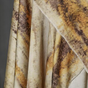 May include: Close-up of a fabric with a marbled pattern in shades of beige, brown, and yellow. The fabric is draped, showing folds and textures. The design resembles natural stone or a watercolor effect.