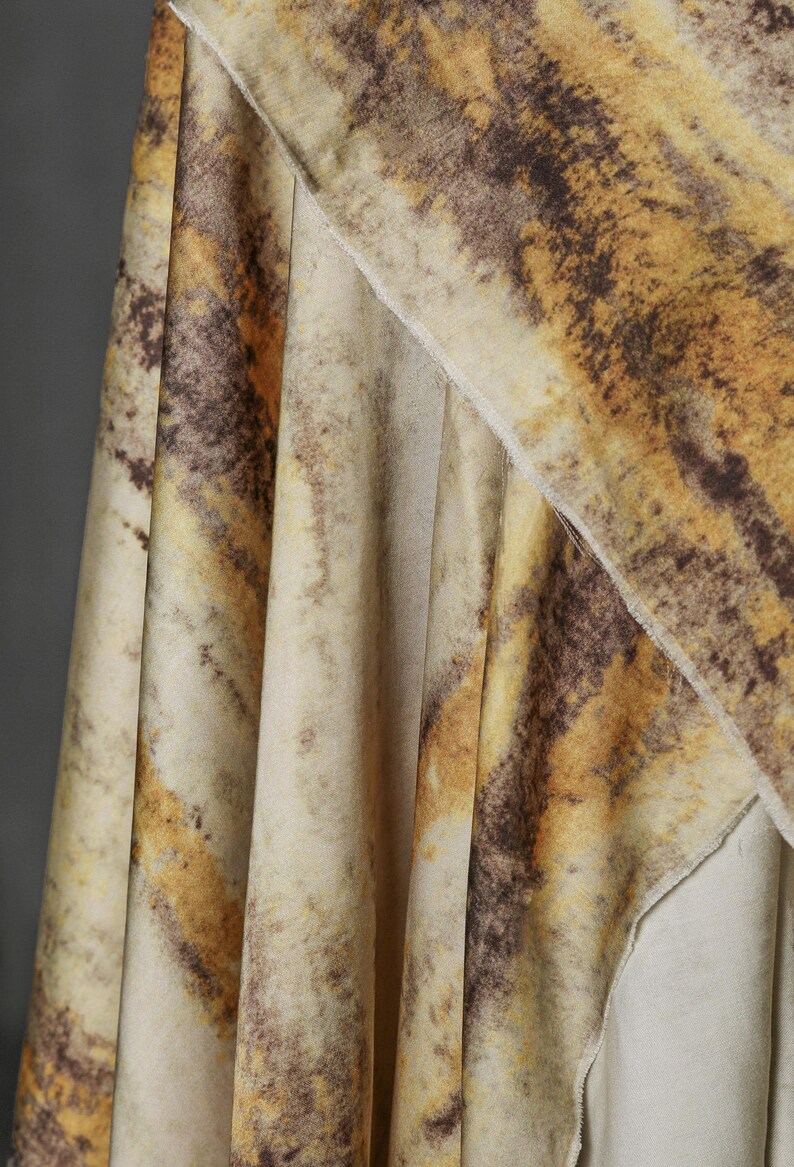 May include: Close-up of a fabric with a marbled pattern in shades of beige, brown, and yellow. The fabric is draped, showing folds and textures. The design resembles natural stone or a watercolor effect.