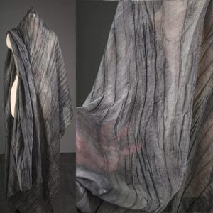 May include: A draped, sheer grey scarf with vertical, textured lines. The fabric is lightweight and appears semi-transparent, with a subtle sheen. The scarf is draped over a mannequin, showcasing its flowing design and potential for layering.