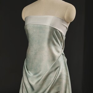 May include: A strapless dress in a soft, flowing fabric, draped elegantly on a mannequin. The dress features a light blue-green color with a contrasting light gray band at the top. The fabric drapes to create a soft, flowing silhouette.