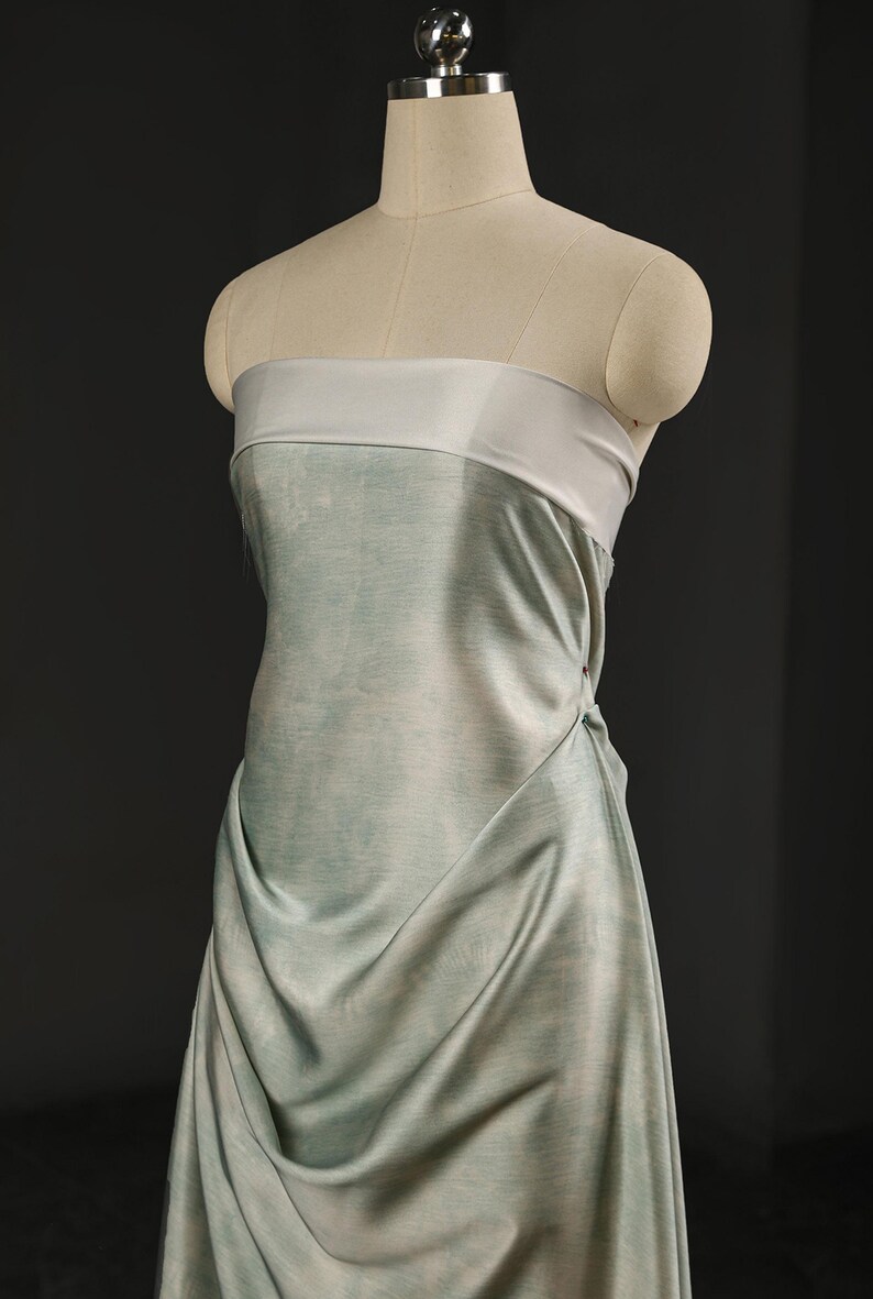 May include: A strapless dress in a soft, flowing fabric, draped elegantly on a mannequin. The dress features a light blue-green color with a contrasting light gray band at the top. The fabric drapes to create a soft, flowing silhouette.