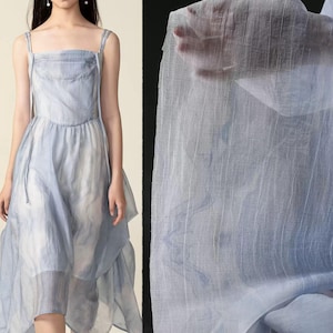 Könnte beinhalten: Ein hellblaues, mehrschichtiges Kleid mit dünnen Trägern. Das Kleid hat einen asymmetrischen Saum und einen halbtransparenten Look. Der Stoff wirkt leicht und luftig, passend für warmes Wetter. Das Kleid ist ein weiches, gedämpftes Blau.