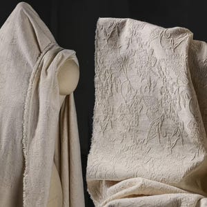 Puede incluir: Tela beige con un patrón floral en relieve. La tela está drapeada sobre un maniquí y también se muestra en una sección doblada, mostrando la textura y el diseño. La tela tiene un borde sin rematar.