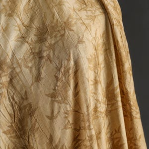 May include: Close-up of a beige fabric with a repeating leaf pattern in a darker shade of beige. The fabric is draped, showing folds and texture. The overall appearance is soft and natural.