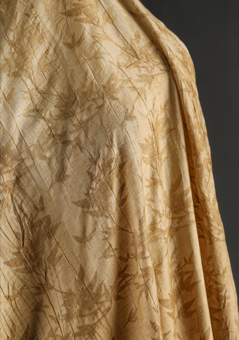 May include: Close-up of a beige fabric with a repeating leaf pattern in a darker shade of beige. The fabric is draped, showing folds and texture. The overall appearance is soft and natural.