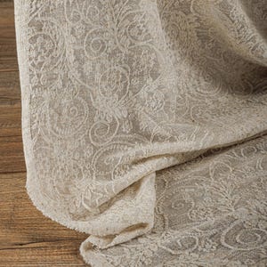 May include: Close-up of a beige fabric with an intricate floral pattern. The fabric is draped, revealing its texture and design. The detailed pattern is visible throughout the cloth, creating a visually rich textile.