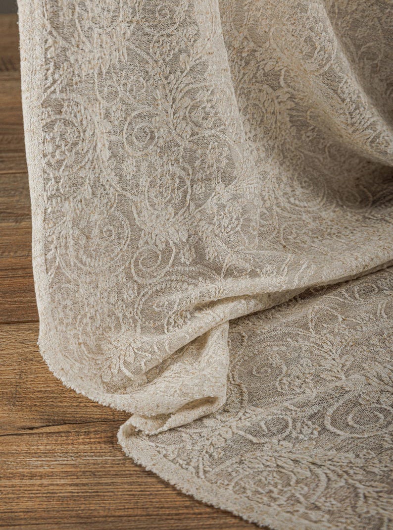 May include: Close-up of a beige fabric with an intricate floral pattern. The fabric is draped, revealing its texture and design. The detailed pattern is visible throughout the cloth, creating a visually rich textile.