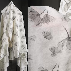 May include: A white scarf featuring a subtle floral print in grey tones. The scarf is draped, highlighting its flowing texture and the intricate botanical design. The fabric appears lightweight and airy, suitable for various styles.