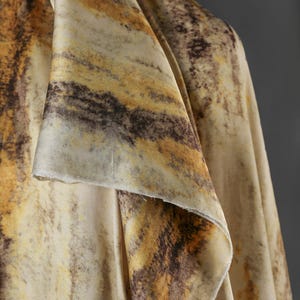 May include: Close-up of a draped fabric with a marbled pattern. The fabric features a blend of colors, including cream, brown, and golden yellow, creating a textured, organic appearance. The edges of the fabric are visible, showing the material's texture.