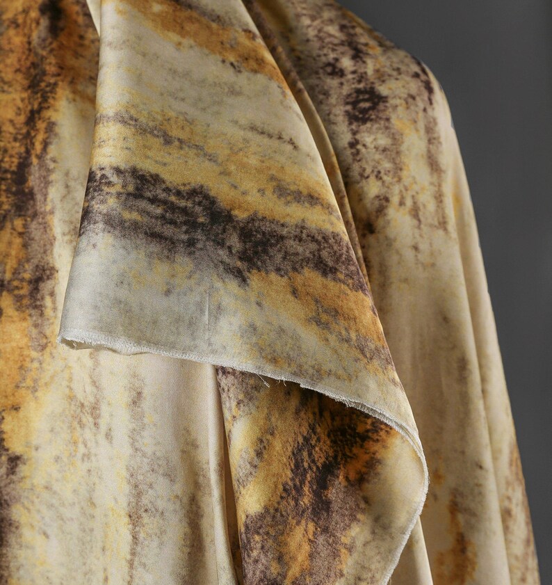 May include: Close-up of a draped fabric with a marbled pattern. The fabric features a blend of colors, including cream, brown, and golden yellow, creating a textured, organic appearance. The edges of the fabric are visible, showing the material's texture.
