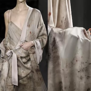May include: A draped, flowing garment in shades of beige and brown, with a subtle pattern and a tie at the waist. The fabric appears to be silk or a similar material, with a soft sheen. The garment is worn over a dress with thin straps.
