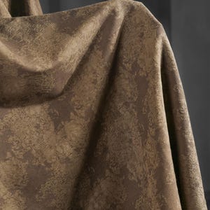 May include: Close-up of a textured fabric in shades of brown and tan. The material has a mottled pattern, with darker brown areas contrasting against lighter tan patches. The fabric is draped, creating folds and shadows.