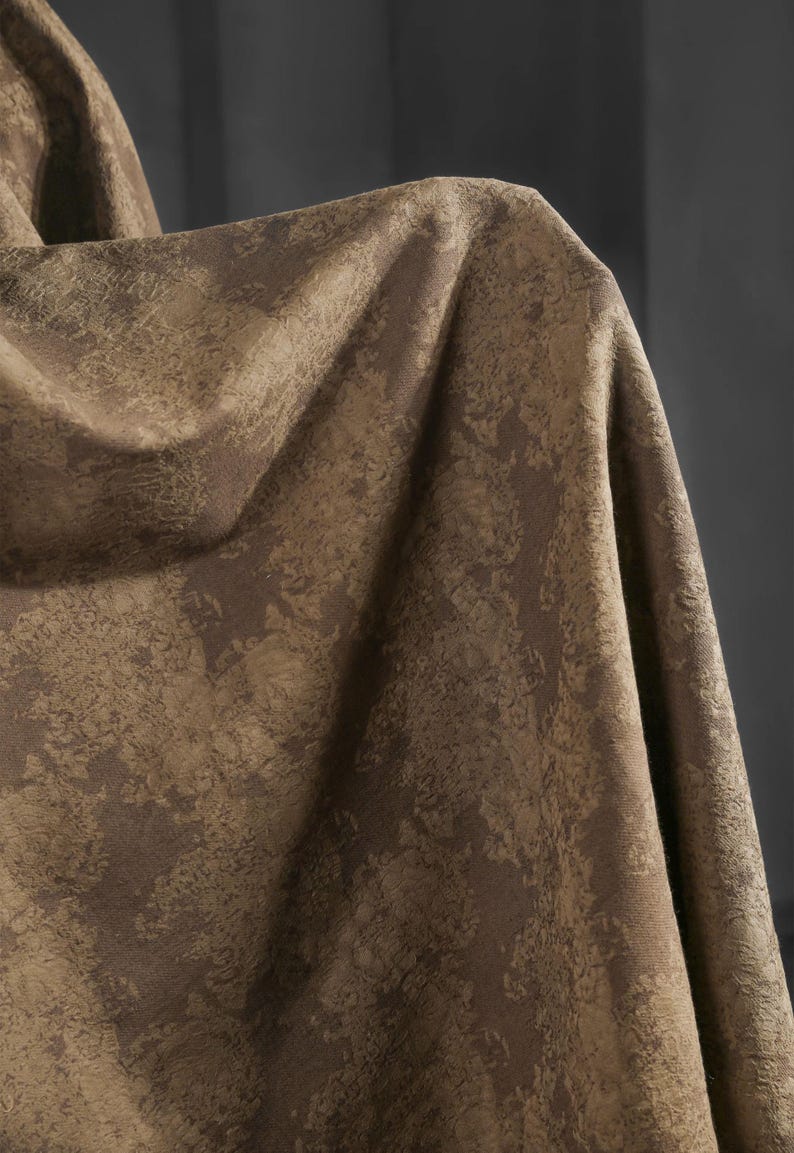 May include: Close-up of a textured fabric in shades of brown and tan. The material has a mottled pattern, with darker brown areas contrasting against lighter tan patches. The fabric is draped, creating folds and shadows.