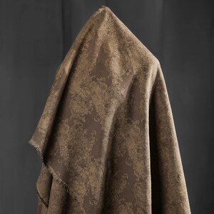 May include: A draped fabric in shades of brown and tan, featuring a textured, mottled pattern. The fabric is folded over a form, creating a draped effect. The edges of the fabric have a fringed detail. The background is a solid, dark gray.