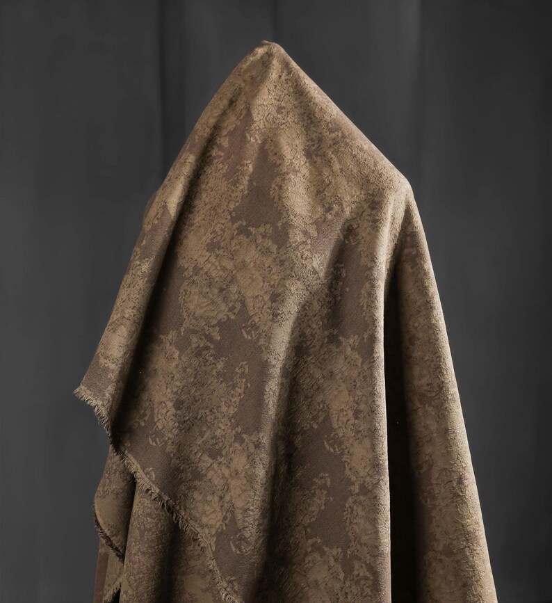 May include: A draped fabric in shades of brown and tan, featuring a textured, mottled pattern. The fabric is folded over a form, creating a draped effect. The edges of the fabric have a fringed detail. The background is a solid, dark gray.