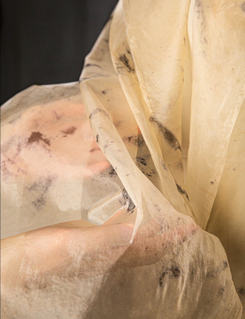 May include: A sheer, cream-colored fabric with dark, irregular markings. The fabric is draped and held, creating soft folds and shadows. The material appears delicate and translucent, with a subtle texture. The background is a dark, solid color.