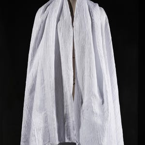 May include: A draped white fabric with a textured, crinkled appearance. The fabric is draped over a mannequin, creating soft folds and shadows. The material appears lightweight and flowing, suitable for clothing or decorative purposes.