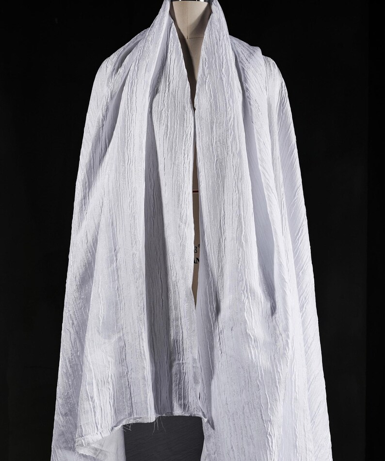 May include: A draped white fabric with a textured, crinkled appearance. The fabric is draped over a mannequin, creating soft folds and shadows. The material appears lightweight and flowing, suitable for clothing or decorative purposes.