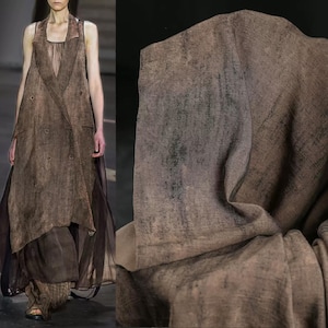 Puede incluir: Un vestido de lino marr&oacute;n con un dise&ntilde;o cruzado y una falda larga y fluida. El vestido tiene un aspecto texturizado y se combina con una superposici&oacute;n transparente de color marr&oacute;n oscuro. La muestra de tela de la derecha muestra un primer plano del material texturizado.