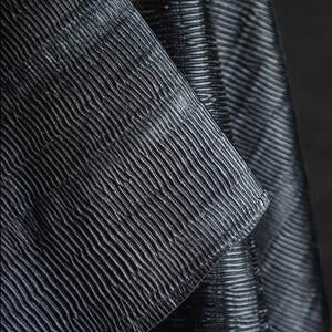 May include: Close-up of a textured fabric with a ribbed pattern. The fabric appears to be dark gray or black, with subtle variations in tone. The ribbed texture creates a play of light and shadow, adding depth to the material.