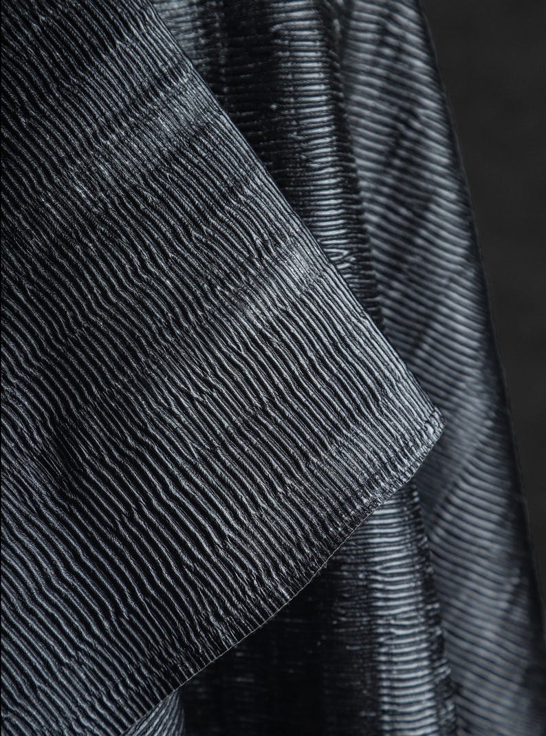 May include: Close-up of a textured fabric with a ribbed pattern. The fabric appears to be dark gray or black, with subtle variations in tone. The ribbed texture creates a play of light and shadow, adding depth to the material.