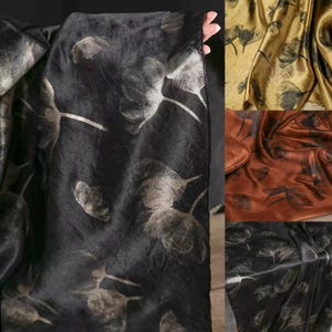 Può includere: Primo piano di un tessuto nero con un motivo floreale grigio e argento. Il tessuto ha una consistenza lucida e setosa. Sono visibili anche campioni di tessuto aggiuntivi in oro e ruggine.