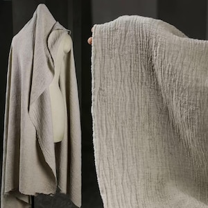 May include: A light beige, textured fabric draped over a mannequin and held up by a hand. The fabric has a crinkled appearance and a raw edge. The fabric is a neutral color, suitable for various textile projects.