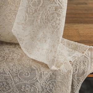 May include: Close-up of a cream-colored, intricately patterned fabric. The fabric is folded over, revealing a detailed floral design. The texture appears soft and slightly textured, suitable for home decor.