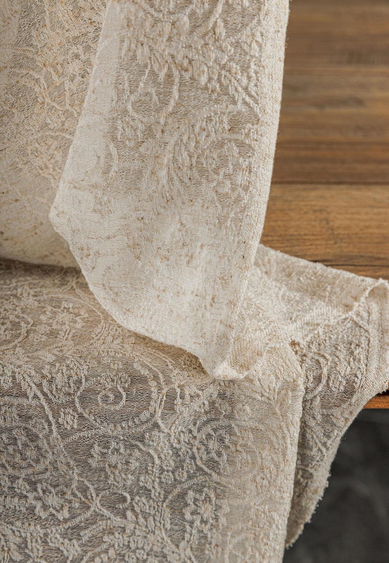 May include: Close-up of a cream-colored, intricately patterned fabric. The fabric is folded over, revealing a detailed floral design. The texture appears soft and slightly textured, suitable for home decor.