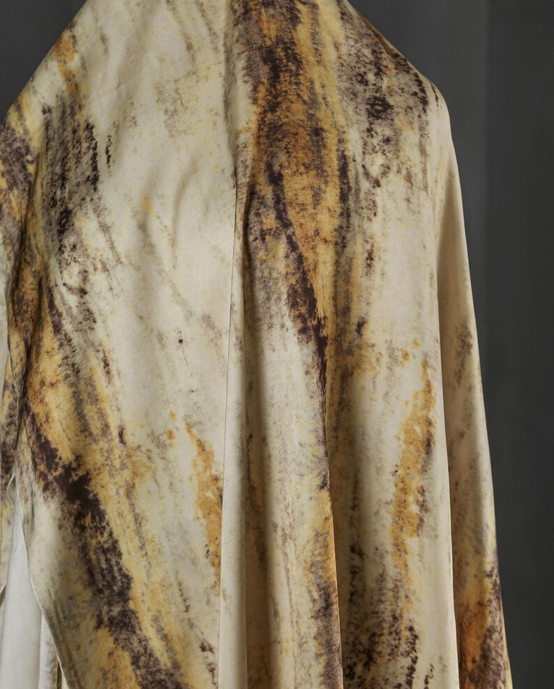 May include: A draped fabric with a marbled pattern. The fabric features a blend of beige, brown, and gold tones, creating a natural, organic aesthetic. The material appears soft and flowing, with visible folds and textures.