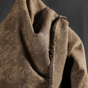 May include: Close-up of a folded brown fabric with a textured, mottled pattern. The fabric has a soft, draped appearance, with visible seams and a fringed edge. The color is a warm, earthy tone.