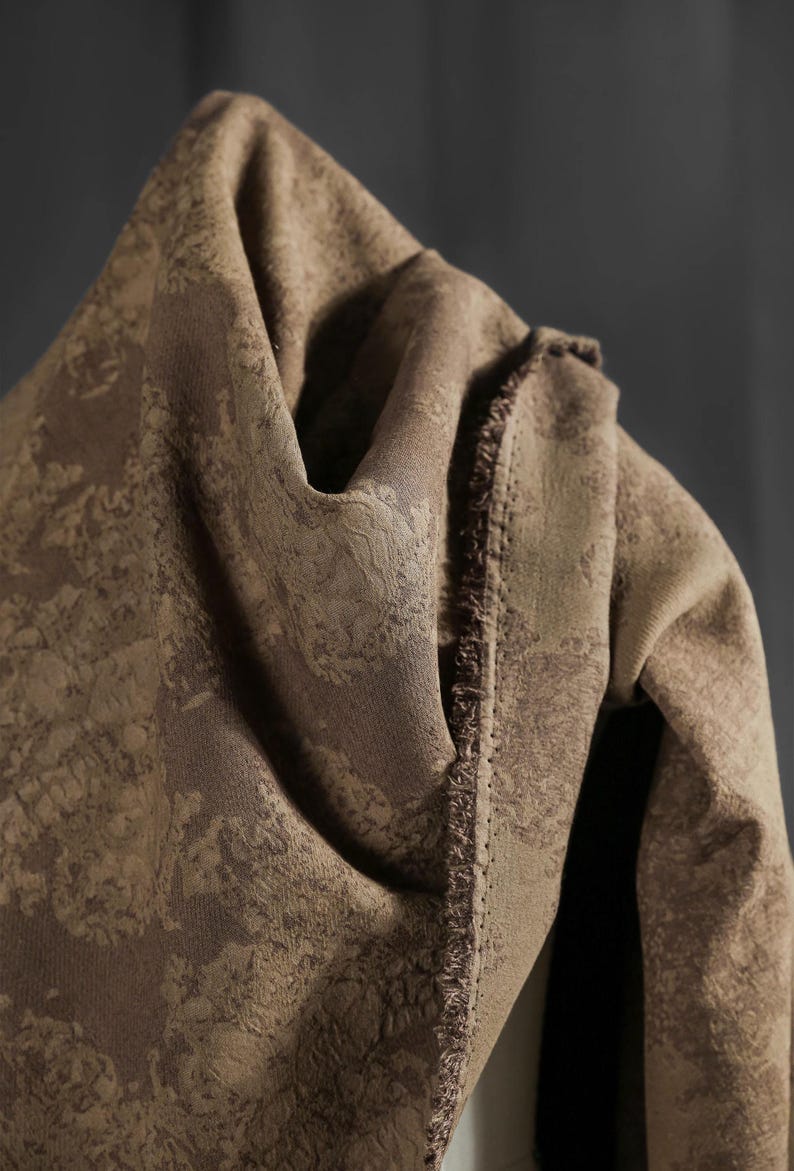 May include: Close-up of a folded brown fabric with a textured, mottled pattern. The fabric has a soft, draped appearance, with visible seams and a fringed edge. The color is a warm, earthy tone.