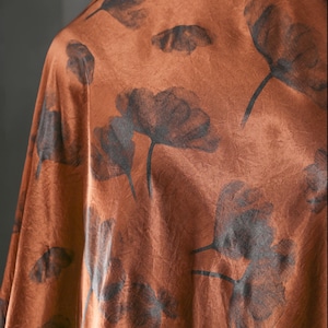 Peut inclure: Gros plan sur un tissu couleur rouille avec un motif floral. Le motif présente des fleurs de pavot stylisées gris foncé avec des tiges. Le tissu a une texture légèrement froissée, suggérant une matière souple.