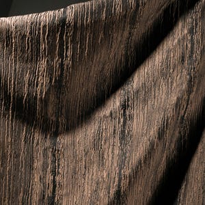 May include: Close-up of a textured fabric with a vertical, linear pattern. The fabric has a dark brown base color with lighter brown, raised lines running from top to bottom. The fabric is draped, creating shadows and highlights.