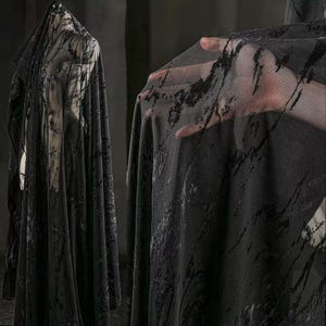 Puede incluir: Tela negra transparente con un patrón oscuro y abstracto. La tela está drapeada sobre un maniquí y sostenida por una mano, mostrando su textura y caída. El patrón parece ser una mezcla de salpicaduras y rayas.