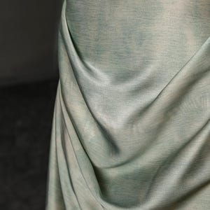 May include: Close-up of a draped fabric in shades of pale green and white. The fabric has a textured appearance, with subtle variations in color and a soft, flowing drape. The folds create shadows and highlights.