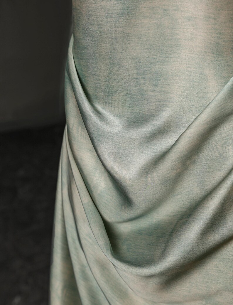 May include: Close-up of a draped fabric in shades of pale green and white. The fabric has a textured appearance, with subtle variations in color and a soft, flowing drape. The folds create shadows and highlights.