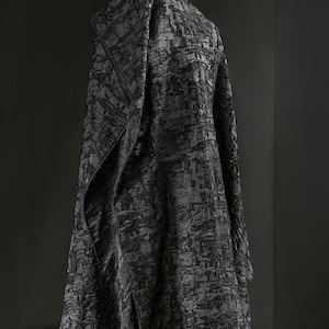 Puede incluir: Una tela gris oscuro, texturizada, drapeada sobre una forma. La tela tiene un patrón de formas rectangulares superpuestas, creando una textura visual. La prenda parece ser un abrigo o chaqueta, con un cuello estructurado.