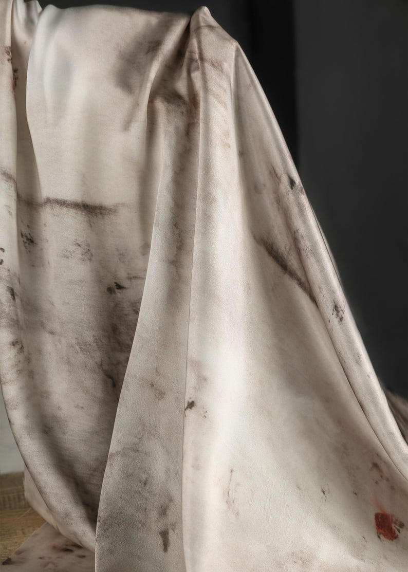 May include: Off-white fabric with dark brown and black mottled patterns. The fabric is draped, showing folds and textures. The overall effect is a blend of light and dark tones, creating a visual contrast. The fabric appears to be lightweight and flowing.