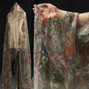 May include: A patterned sheer fabric in shades of green, red, and brown, draped over a mannequin. The fabric is held up by a hand, highlighting its flowing texture. The design features an abstract pattern, reminiscent of a painting.