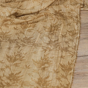May include: Tan fabric with a repeating pattern of brown leaves and stems. The fabric has a textured, pleated appearance, and is draped over a wooden surface. The edges of the fabric are frayed.
