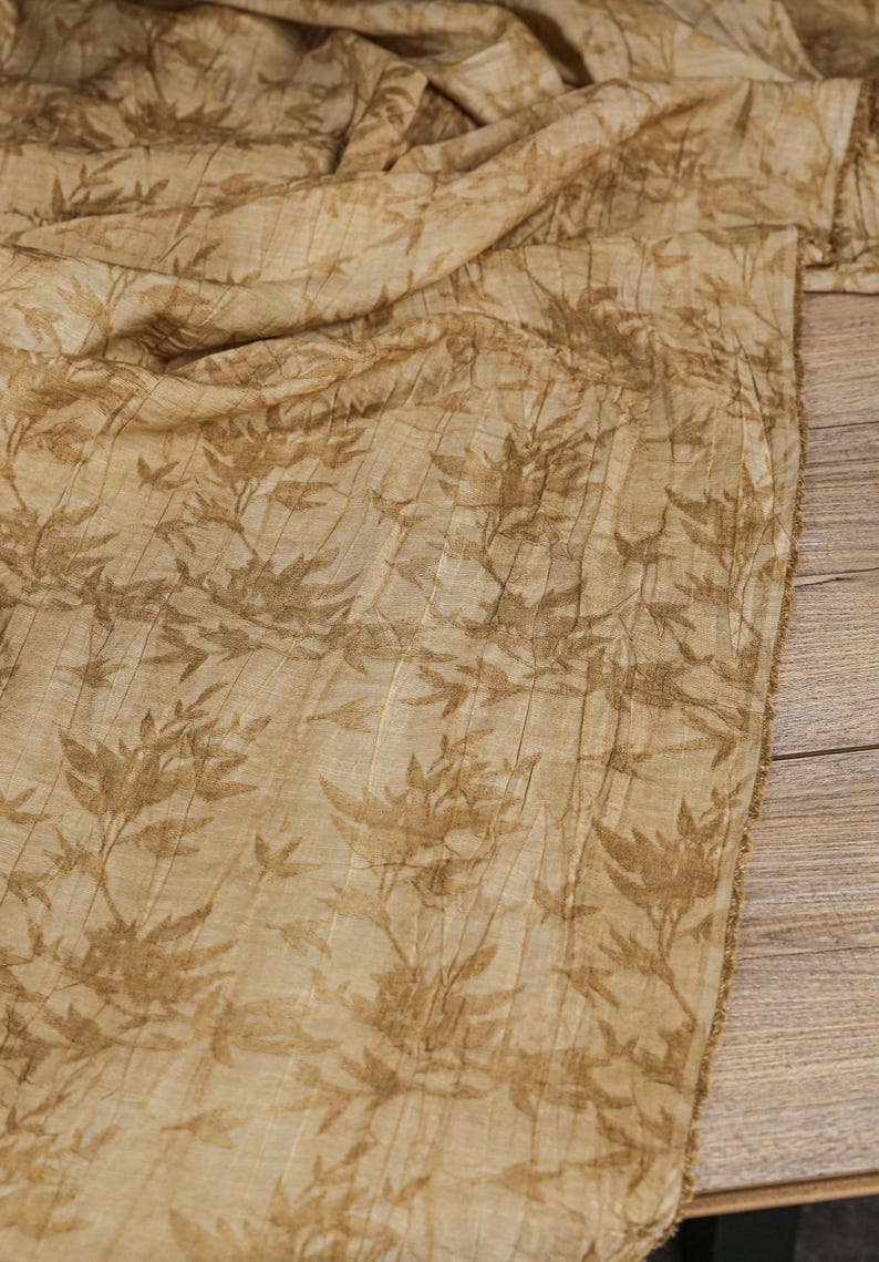 May include: Tan fabric with a repeating pattern of brown leaves and stems. The fabric has a textured, pleated appearance, and is draped over a wooden surface. The edges of the fabric are frayed.