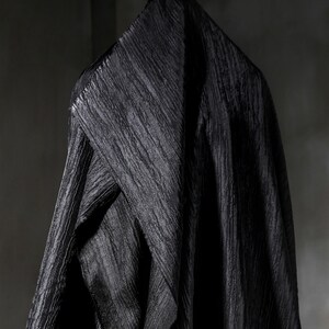May include: A draped black fabric with a textured, rippled surface hangs against a dark gray background. The fabric is folded over a hanger, creating a triangular shape at the top. The material appears to be heavy and opaque.