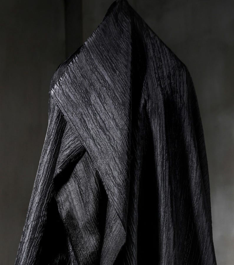May include: A draped black fabric with a textured, rippled surface hangs against a dark gray background. The fabric is folded over a hanger, creating a triangular shape at the top. The material appears to be heavy and opaque.