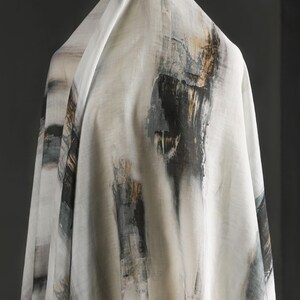 May include: White fabric with an abstract design featuring vertical brushstrokes in black, grey, and gold. The fabric is draped, showcasing the pattern and texture. The overall aesthetic is artistic and modern, suitable for apparel or home decor.