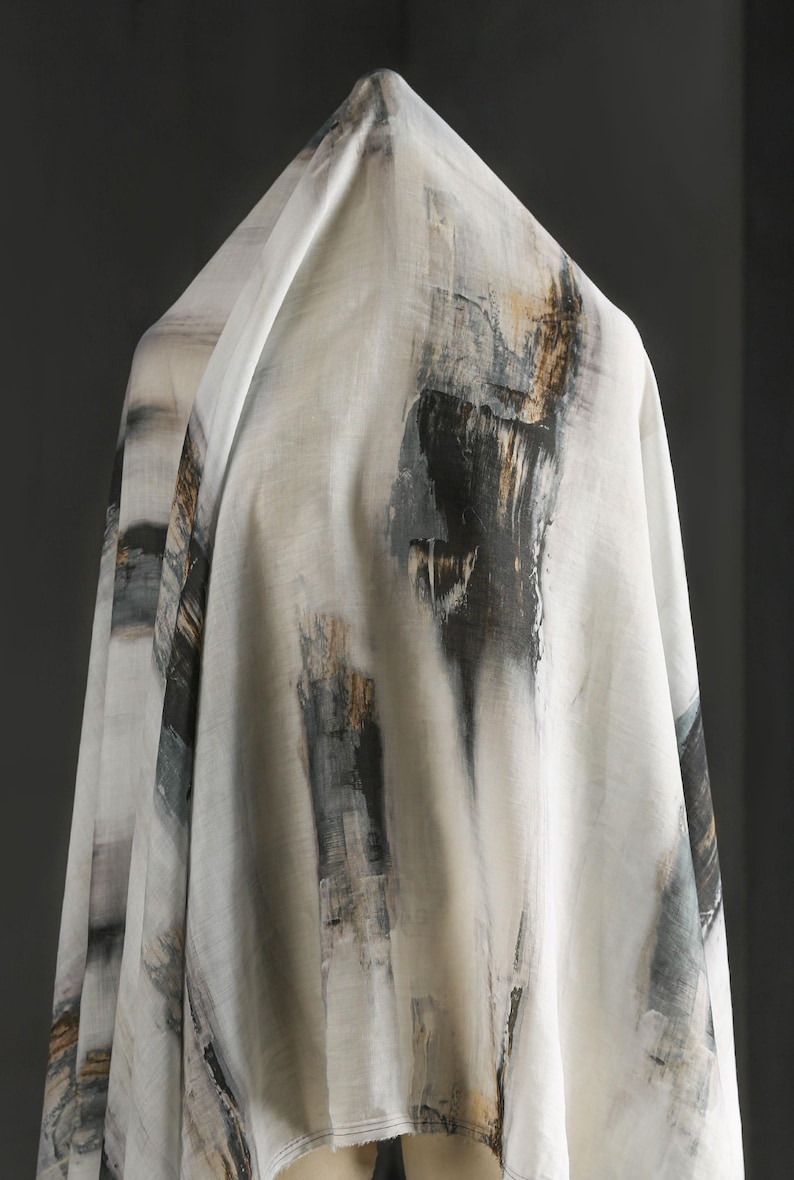 May include: White fabric with an abstract design featuring vertical brushstrokes in black, grey, and gold. The fabric is draped, showcasing the pattern and texture. The overall aesthetic is artistic and modern, suitable for apparel or home decor.
